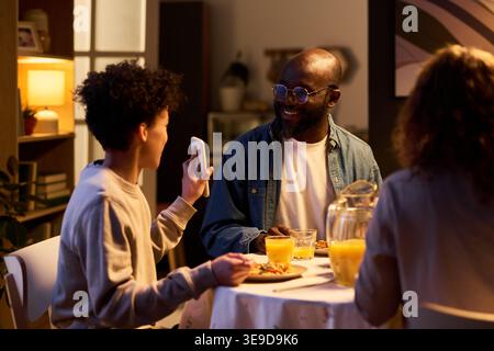 Padre afroamericano sorridente all'adolescente che mostra lo smartphone durante la cena di famiglia, discutendo di contenuti digitali e connessione. Ideale per i genitori, lo schermo e il marketing per lo stile di vita familiare Foto Stock