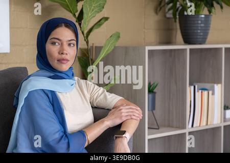 Donna che indossa un hijab blu seduta su una poltrona grigio scuro in salotto con smartwatch Foto Stock