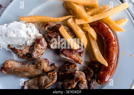 Piatto greco tradizionale con pezzi di maiale alla griglia, salsiccia abbondante, patatine fritte dorate e salsa tzatziki cremosa su un piatto bianco Foto Stock