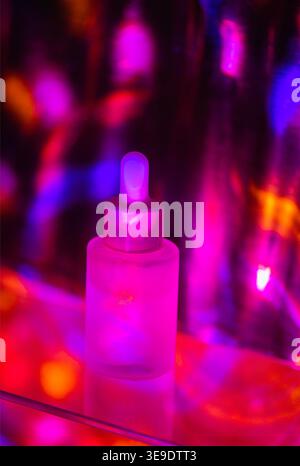 Flacone contagocce trasparente con tappo bianco illuminato da luci colorate, su sfondo sfocato con vivaci colori rosa, viola e blu Foto Stock