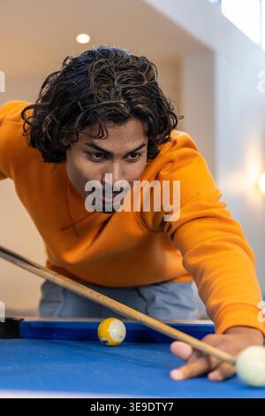 Uomo indiano degli anni Venti che indossa un maglione arancione appoggiato sul tavolo da biliardo in feltro blu a casa, puntando il segno. Tavoli da biliardo, concentrazione, ambiente, illuminazione, lucernario, tempo libero, hobby Foto Stock