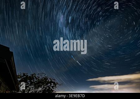 Composito a lunga esposizione che mostra percorsi stellari circolari centrati su un palo celeste meridionale sopra un albero sagomato e un edificio, con satelliti luminosi Foto Stock