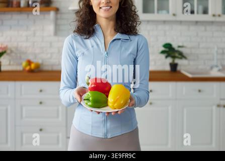 Donna sorridente che tiene il piatto in piedi nella cucina moderna di casa, tre vivaci peperoni Foto Stock