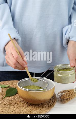 Donna setacciando polvere di matcha nel recipiente al tavolo bianco, primo piano Foto Stock