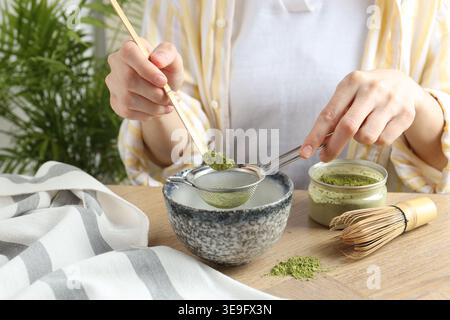 Donna setacciando polvere di matcha nel recipiente al tavolo di legno, primo piano Foto Stock
