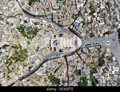 Vista dall'alto con drone della Chiesa di San Nicola a Siggiewi, circondata da tetti di pietra calcarea, stradine strette, auto parcheggiate e una piccola piazza. Foto Stock