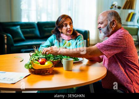 Una coppia indiana anziana seduta insieme a un tavolo per la colazione e facendo una piacevole conversazione mentre entrambi stanno attivamente smistando verdure verdi fresche Foto Stock