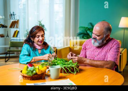 Una coppia indiana anziana seduta insieme a un tavolo per la colazione e facendo una piacevole conversazione mentre entrambi stanno attivamente smistando verdure verdi fresche Foto Stock