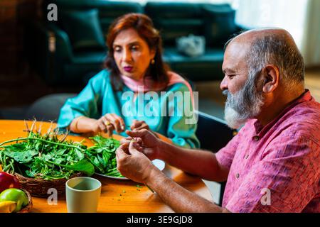 Una coppia indiana anziana seduta insieme a un tavolo per la colazione e facendo una piacevole conversazione mentre entrambi stanno attivamente smistando verdure verdi fresche Foto Stock