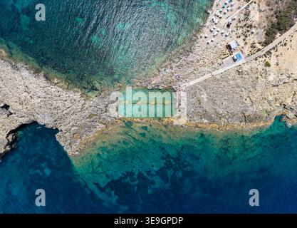 Vista aerea della Qawra Pool, una piscina costiera scavata nella roccia sulla costa mediterranea, con acqua turchese, rocce calcaree e ombrelloni Foto Stock