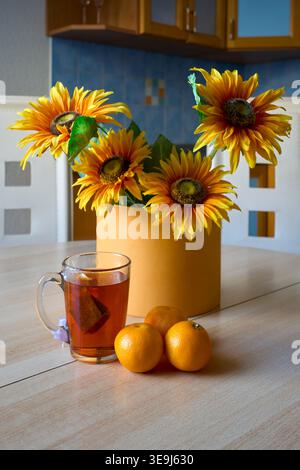 Luminosi girasoli in un vaso giallo accanto a un bicchiere di tè e arance fresche su un tavolo bianco. Atmosfera familiare calda e intima Foto Stock