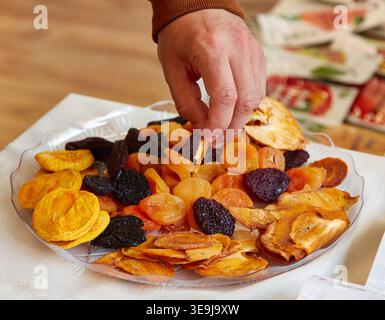 Selezione a mano di vari tipi di frutta secca da un piatto trasparente, tra cui albicocche, prugne e fette di mele, disposte su una tovaglia bianca in un ambiente accogliente Foto Stock