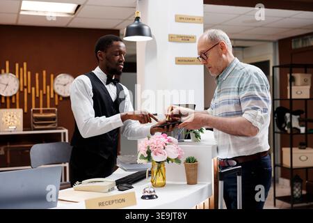Un anziano maturo in piedi al banco della reception dell'hotel, dove un cortese addetto alla reception aiuta a prenotare su un tablet digitale. Un cliente maschio anziano firma un modulo di check-in online sul dispositivo smart. Foto Stock