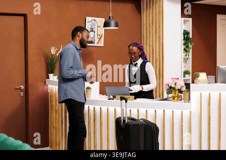 La receptionist afroamericana sorridente e cordiale parla con gli ospiti dell'hotel alla reception, controllando il passaporto. Viaggiatore nero in piedi con la valigia alla reception durante la procedura di check-in Foto Stock