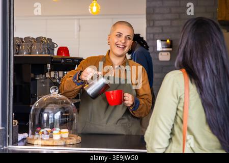 Barista in grembiule verde oliva che versa il latte dalla caraffa metallica nella tazza rossa, il cliente tiene in mano il sacchetto Foto Stock