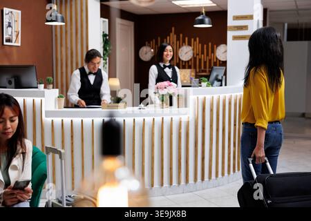 I vacanzieri arrivano all'invitante ingresso dell'hotel con i bagagli e aspettano di prenotare la camera. Elegante lobby dell'hotel con ospiti freschi e due receptionist asiatici sorridenti Foto Stock