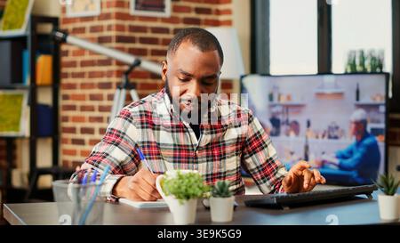 Multitasking, freelance stanchi, controllo incrociato delle informazioni, trascrizione dei dati dallo schermo del computer al blocco note. Telelavoratore afroamericano concentrato che lavora a distanza in una casa accogliente ed elegante, da vicino Foto Stock