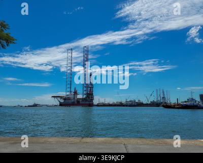 Il grande carro di perforazione offshore galleggia in acque blu calme. Il cielo è chiaro con nuvole bianche sparse sopra. Navi e barche distanti costellano l'orizzonte del porto Foto Stock