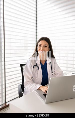 Medico che guarda la macchina fotografica, seduto alla scrivania con il computer portatile Foto Stock