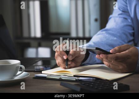 Tenere a mano lo smartphone mentre si scrivono appunti sul notebook accanto al laptop, alla scheda, alla calcolatrice e alla tazza di caffè sulla scrivania, mostrando la pianificazione dei contenuti e la risoluzione digitale Foto Stock