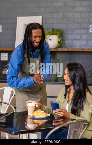 Server diversificato in grembiule verde appoggiato, sorridente, cliente donna che tiene il telefono e porta la tazza al bar. Barista, dreadlocks, denim, croissant, pasticceria, caffetteria, brick Foto Stock