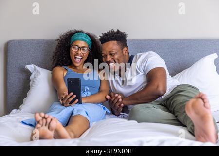 Diverse coppie sdraiate sul letto ridendo tenendo in mano lo smartphone nero, donna con un abito blu. Camera da letto, intimità, romanticismo, telefono, relax, naturale, caldo Foto Stock