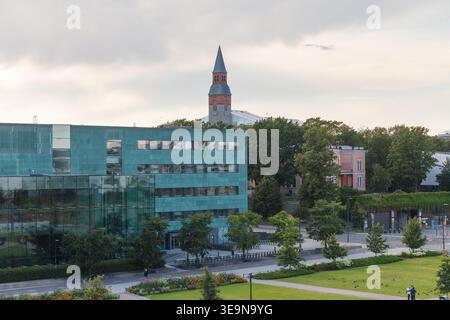 HELSINKI, FINLANDIA - 11 AGOSTO 2025: Edificio per uffici in vetro turchese con una torre in mattoni rossi che si innalza sopra alberi verdi in estate. Foto Stock