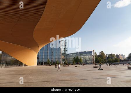 HELSINKI, FINLANDIA - 11 AGOSTO 2025: Ampia piazza pubblica con un baldacchino dorato curvo e un edificio a torre di vetro sotto un cielo estivo. Foto Stock