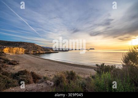 Alba mozzafiato a la Carolina Beach a Águilas. Il sole sorge all'orizzonte e si riflette sul mare calmo, mentre la vegetazione costiera è al foregro Foto Stock