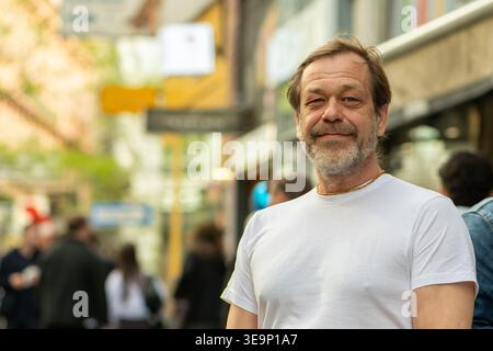 Uomo adulto di 50 anni che posa per strada in città in un abbigliamento casual con pedoni sullo sfondo. Foto Stock