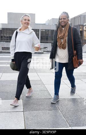 Amici di sesso femminile che camminano attraverso la piazza fuori dall'edificio civico, tenendo tazze da caffè Foto Stock