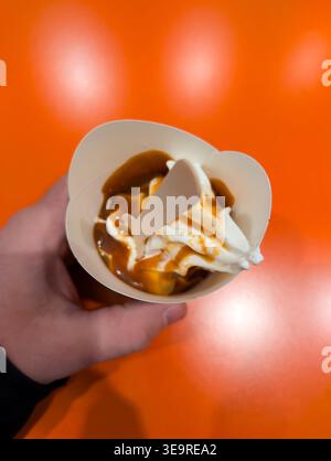 Vista dall'alto del gelato alla vaniglia servito con salsa al caramello in una tazza di carta con un cucchiaio di legno tenuto a mano. Foto Stock