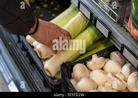 Mano che tiene porri freschi sopra il cestino dei prodotti, preparatore di pasti che controlla la croccantezza, gambi verdi e basi bianche, Bright Market Aisle Closeup Foto Stock