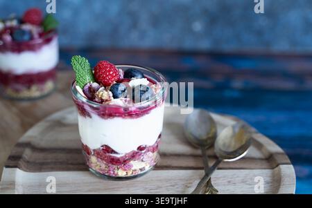Vista frontale dello yogurt parfait con frutti di bosco, cereali e crocchette di biscotti su tavola di legno con sfondo rustico blu Foto Stock
