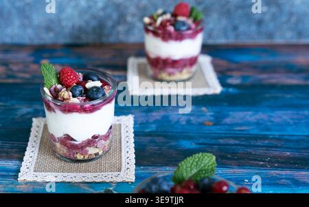 Vista frontale del dessert con yogurt a strati con cereali di frutti di bosco e briciole di biscotti su una superficie di legno blu con spazio per le copie Foto Stock