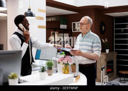 Un anziano ragazzo caucasico in pensione firma i documenti di prenotazione al banco della hall dell'hotel, mentre un dipendente afroamericano riceve una chiamata. Il cliente senior compila i documenti per il check-in mentre l'addetto alla reception è al telefono. Foto Stock