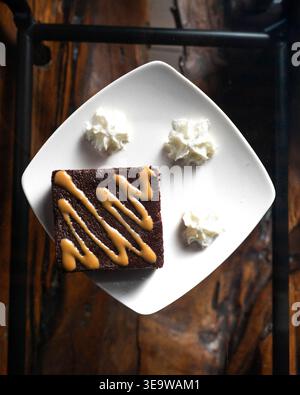 Il piatto bianco quadrato contiene un ricco brownie al cioccolato. Il caramello lancia zigzag sulla sua superficie scura e umida. Tre panna montata circondano il de Foto Stock