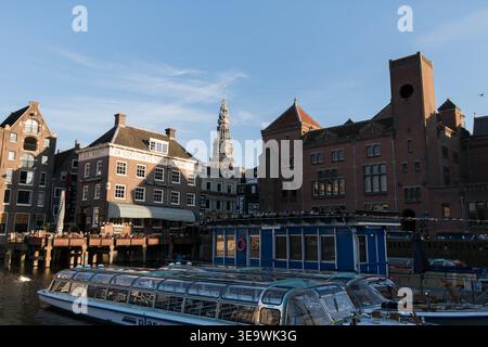Barche da crociera con tetto in vetro ormeggiate sul Damrak ad Amsterdam, con storiche case sui canali e la torre Munttoren sullo sfondo. Foto Stock
