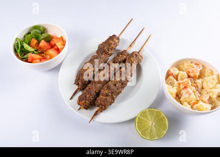 Tre spiedini di kebab lula grigliati si trovano su un piatto bianco con insalate e fette di lime. La presentazione del cibo mostra texture e colori sotto una luminosa illuminazione da studio. Foto Stock