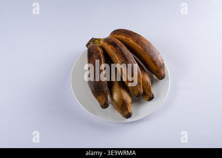 Piantare banane su piatto bianco, con sfumature da marrone a giallo. Illuminazione soffusa e diffusa. Concetto di cibo semplice. Il contrasto testuale aggiunge profondità. Focu Foto Stock