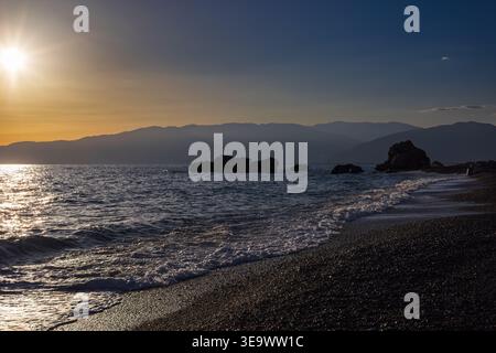 Tramonto sulla spiaggia di ciottoli con montagne e rocce sagomate. Foto Stock