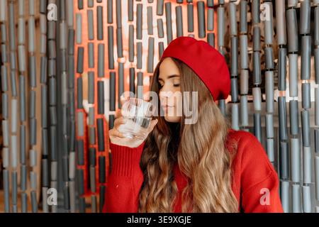 Donna Beret acqua che beve da un bicchiere in un caffè mentre indossa un maglione rosso per una sana idratazione Foto Stock