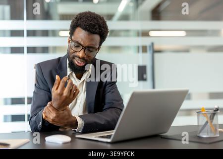 Uomo d'affari alla scrivania dell'ufficio che tiene il polso in difficoltà mentre lavora su un notebook, suggerendo sforzi ripetitivi, rischi nel tunnel carpale e necessità di soluzioni ergonomiche sul posto di lavoro Foto Stock