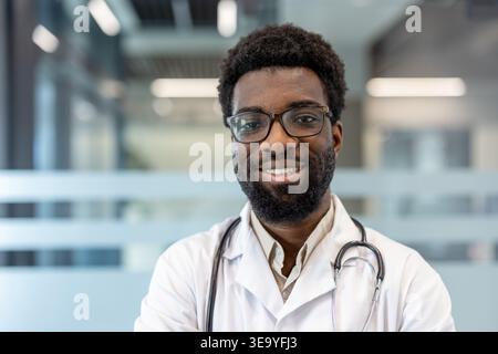 Medico professionista maschile che guarda la fotocamera e sorride, indossa un camice da laboratorio bianco e uno stetoscopio, posare contro un moderno background clinico, che rappresenta l'esperienza sanitaria e la cura del paziente Foto Stock