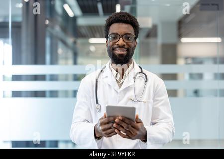 Medico afroamericano con stetoscopio, che sorride e tiene in mano una compressa in un ambiente clinico moderno, simboleggiando l'assistenza sanitaria digitale e l'esperienza focalizzata sul paziente Foto Stock