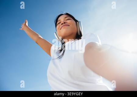 Una giovane donna sta all'aperto sotto un cielo blu limpido con le cuffie indossate, le braccia allungate mentre ascolta la musica, sperimentando libertà e gioia in una p Foto Stock