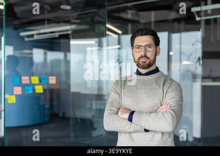 Uomo d'affari con il barbuto che indossa occhiali e un maglione, in piedi con le braccia incrociate, rappresentando la leadership e la professionalità in un ambiente d'ufficio moderno e luminoso Foto Stock