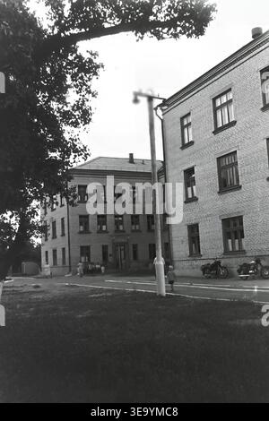 Sloviansk, 1956-1960, via Shevchenko. Questa storica fotografia d'archivio mostra la robusta architettura in mattoni a più piani dell'ospedale Lenin. Diversi individui, probabilmente personale medico e visitatori, sono riuniti vicino all'ingresso principale sotto l'ombra di alberi maturi. Le motociclette d'epoca con sidecar sono parcheggiate ordinatamente lungo il vialetto asfaltato. L'edificio presenta numerose grandi finestre, tetti piastrellati ed elementi strutturali classici che mostrano il sistema sanitario del tranquillo Donbas. Foto Stock