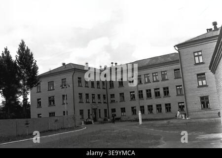Sloviansk, 1956-1960, via Shevchenko. Questa storica fotografia d'archivio mostra la robusta architettura in mattoni a più piani dell'ospedale Lenin. Diversi individui, probabilmente personale medico e visitatori, sono riuniti vicino all'ingresso principale sotto l'ombra di alberi maturi. Le motociclette d'epoca con sidecar sono parcheggiate ordinatamente lungo il vialetto asfaltato. L'edificio presenta numerose grandi finestre, tetti piastrellati ed elementi strutturali classici che mostrano il sistema sanitario del tranquillo Donbas. Foto Stock