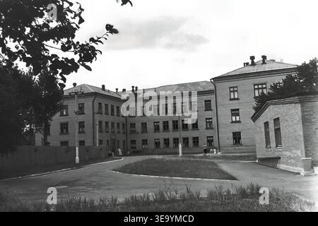 Sloviansk, 1956-1960, via Shevchenko. Questa storica fotografia d'archivio mostra la robusta architettura in mattoni a più piani dell'ospedale Lenin. Diversi individui, probabilmente personale medico e visitatori, sono riuniti vicino all'ingresso principale sotto l'ombra di alberi maturi. Le motociclette d'epoca con sidecar sono parcheggiate ordinatamente lungo il vialetto asfaltato. L'edificio presenta numerose grandi finestre, tetti piastrellati ed elementi strutturali classici che mostrano il sistema sanitario del tranquillo Donbas. Foto Stock